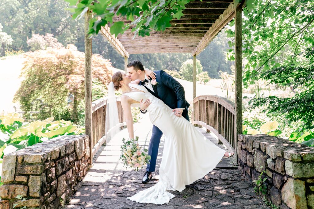 A bride and groom share a dramatic dip kiss in front of a wooden bridge