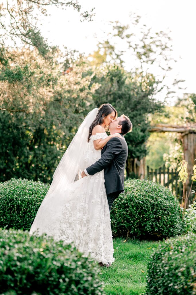 A bride and groom share a kiss as he lifts her off her feet in the garden at golden hour