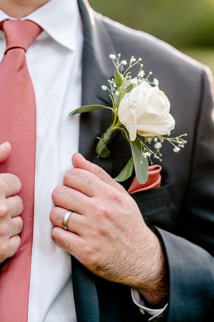 A close up of a groom's boutonnière and tie and ring on his finger after a St. John the Beloved wedding