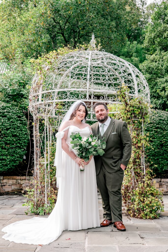 A bride and groom smiling for a classic portrait in front of a white pergola covered in greenery