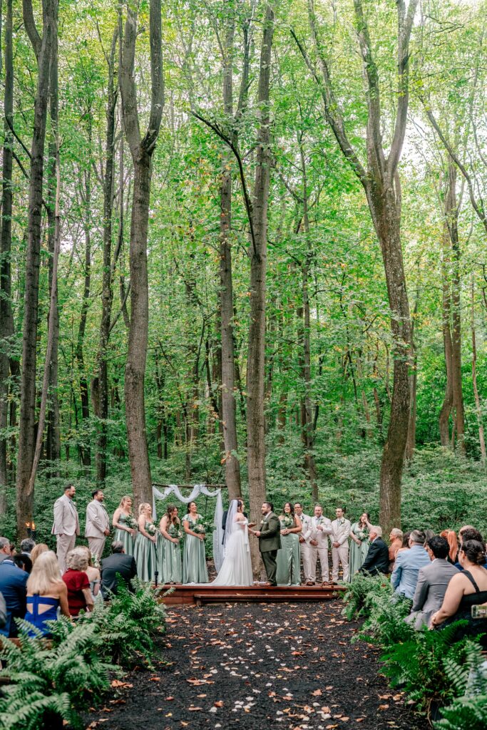 An outdoor wedding ceremony in the middle of woods with tall trees and ferns lining the aisle