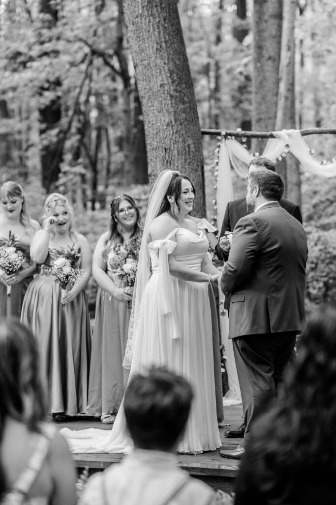 A bride smiling at her groom as a bridesmaid wipes a tear from her eye during an outdoor wedding ceremony