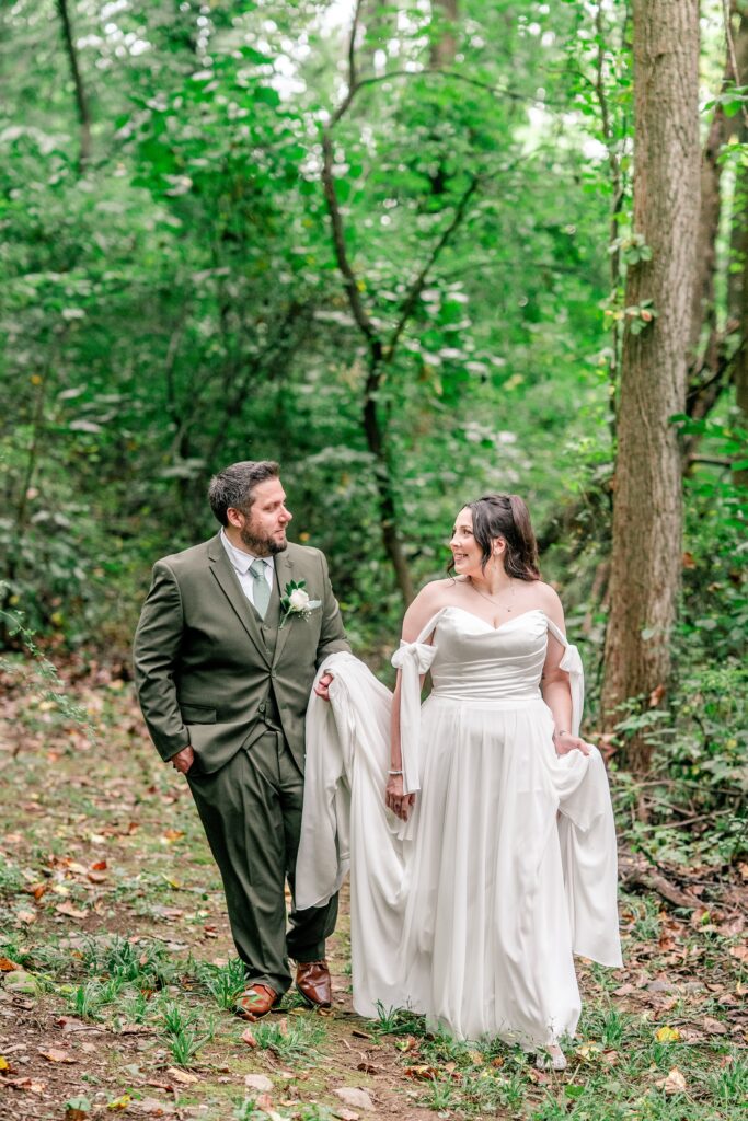 A bride and groom walking together through the woods as he holds her train toward their Northern VA wedding photographer