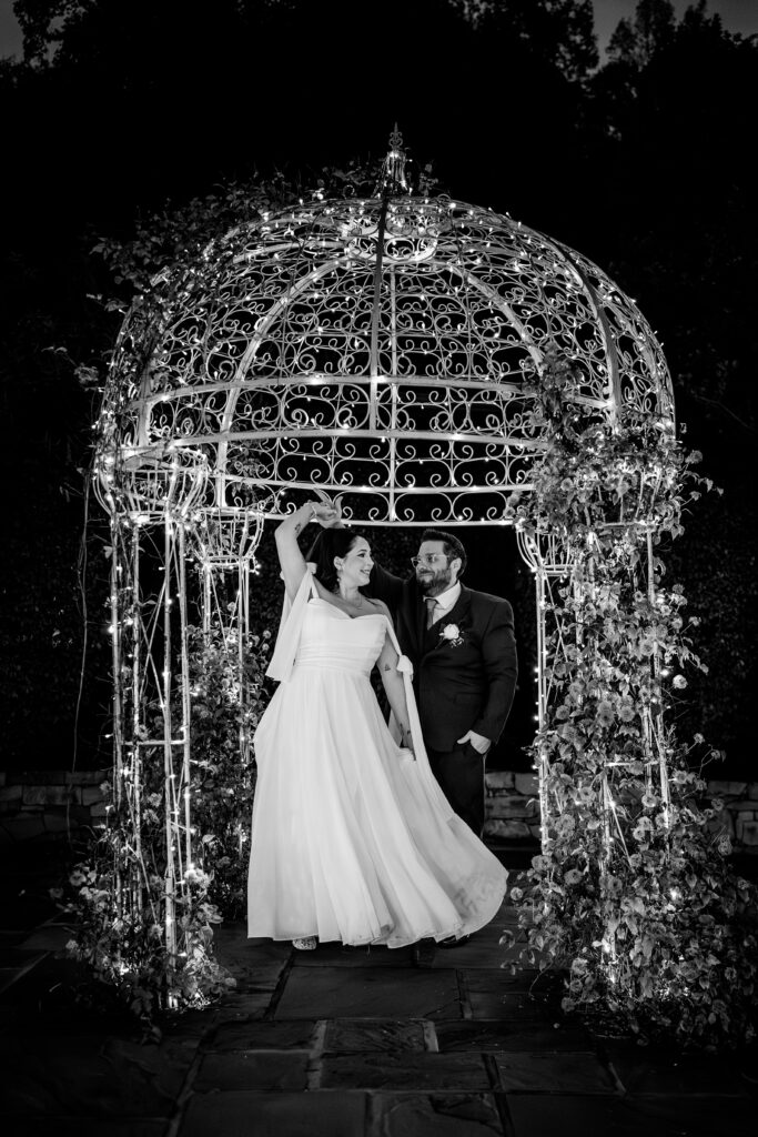 A bride and groom share a dance under a white pergola with string lights during night portraits
