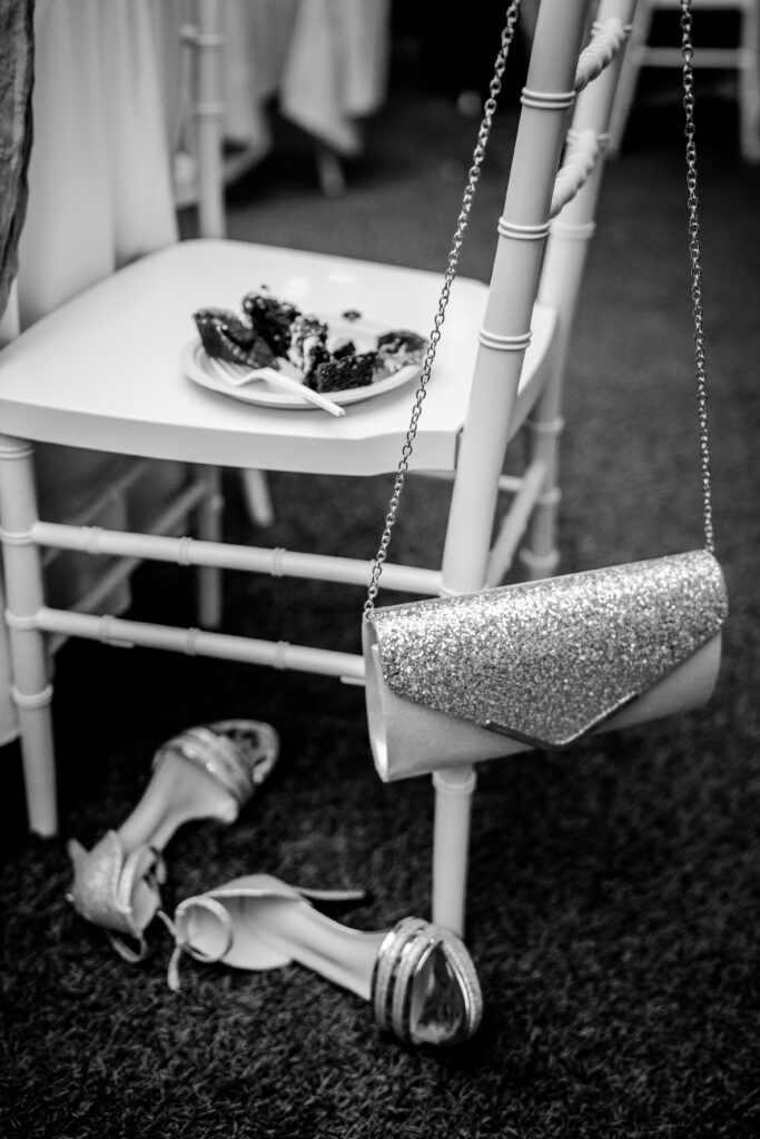 A pair of silver heels sitting below a Stone Manor Boutique Inn wedding reception chair with a sparkling purse draped over the back and a plate of cake on the seat