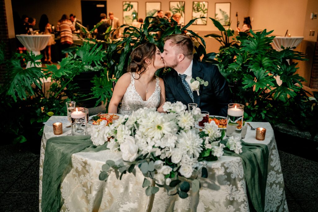 A bride and groom share a kiss at their sweetheart table surrounded by greenery