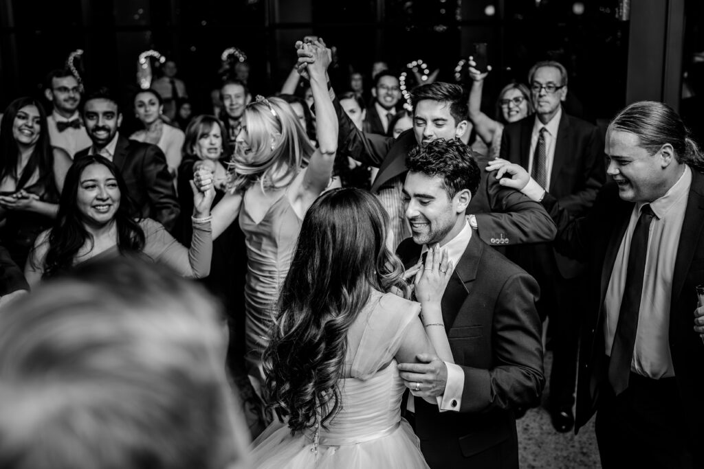 A bride and groom smile at one another as guests cheer on the dance floor