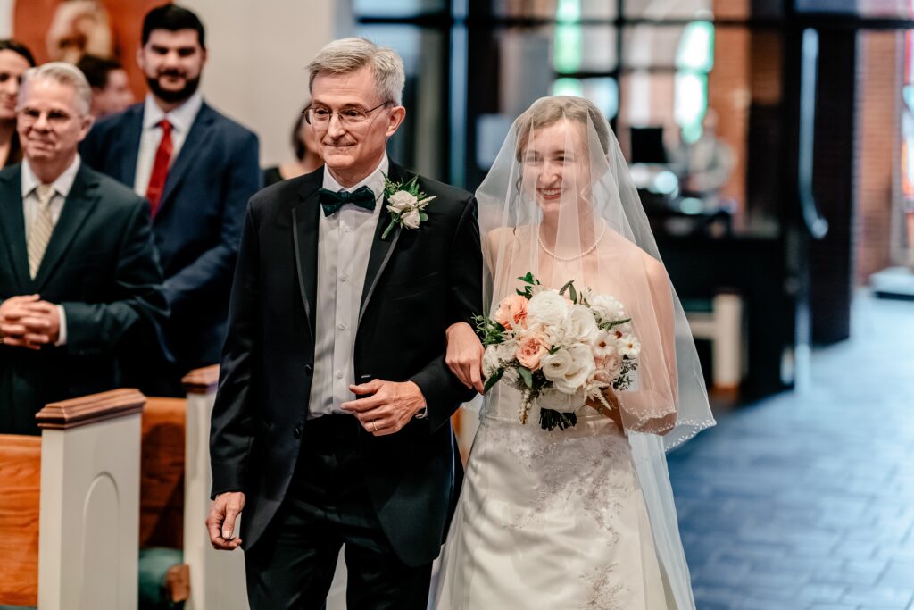 A bride smiles at her groom as she walks down the aisle with her father