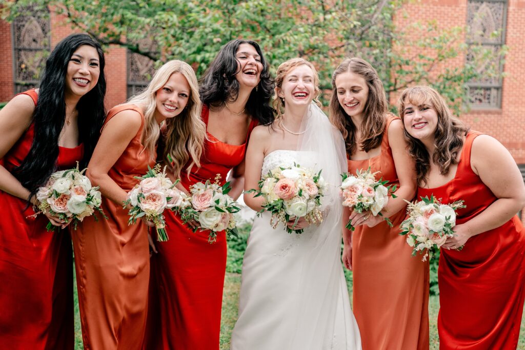 Bridesmaids laugh together wearing orange and peach dresses and carrying pink and peach bouquets