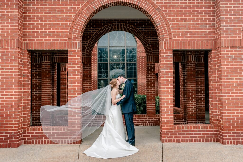 A bride and groom kiss outside their church with her veil flying behind her