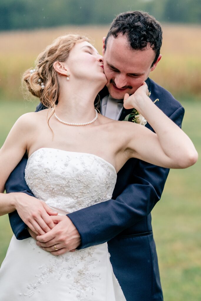A bride kisses her groom on the cheek as he wraps his arms around her wais