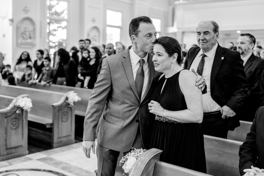 The father of the bride kisses the top of his wife's head after handing off the bride to the groom during their St Francis de Sales wedding in Purcellville