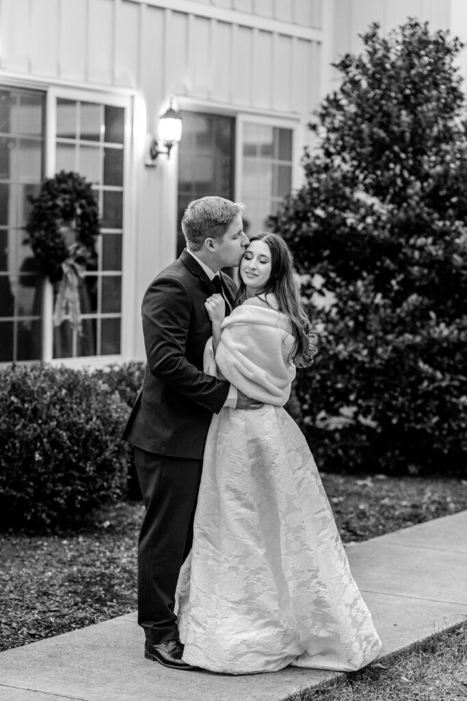 A groom kissing his bride on the head while posed outside the barn for their Historic Rosemont wedding