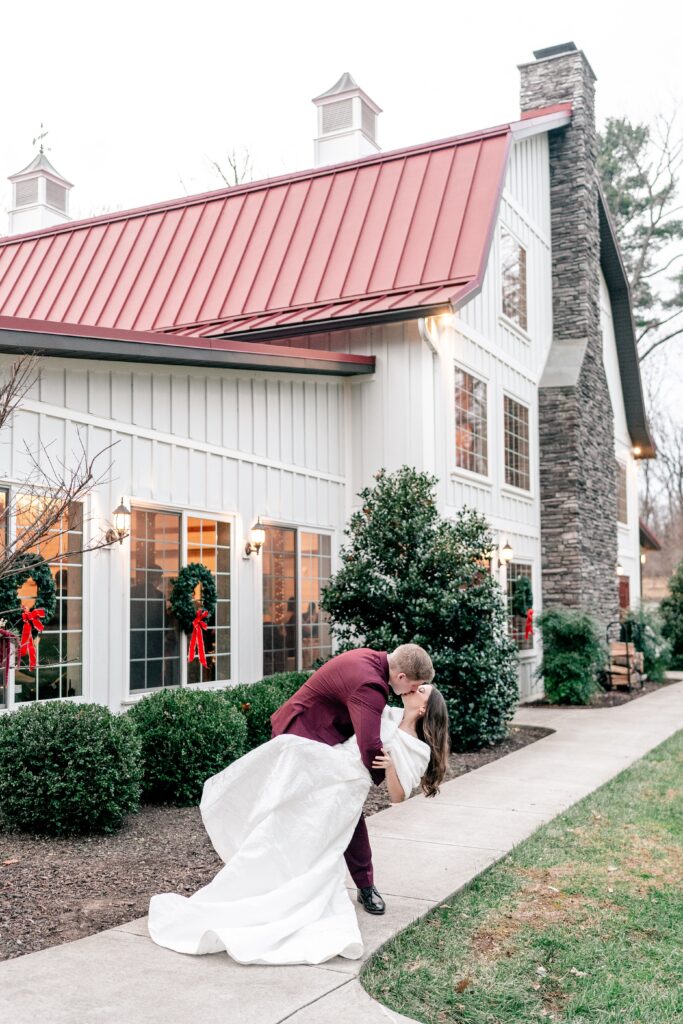 A bride and groom share a dramatic dip kiss outside the barn decorated for Christmas during their Historic Rosemont wedding