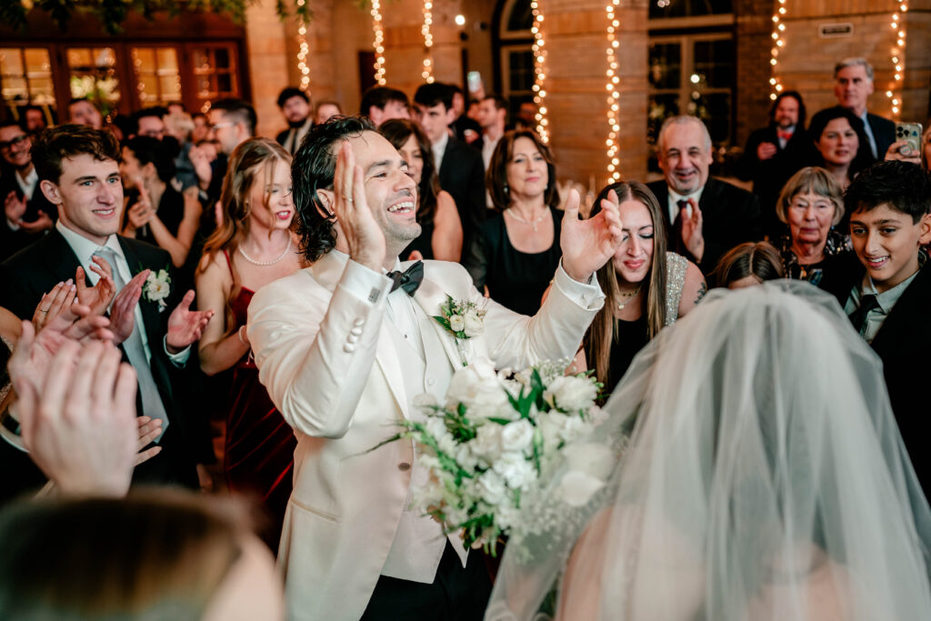 A groom laughing as he claps his hands surrounded by guests on the dance floor