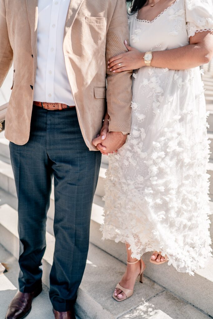 A close up of an engaged couples' outfits as they walk down a set of stairs, featuring a khaki jacket and slacks on the man, and a white floral dress and gold accessories on the woman