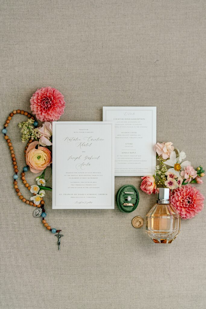 A flat lay of wedding day details including invitations, a rosary, perfume, and rings