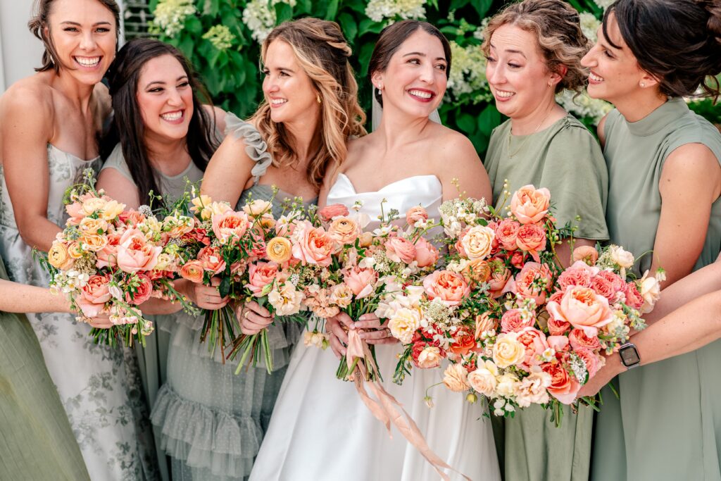 A bride smiling at her bridesmaids as they hold summer wedding bouquets with peach flowers