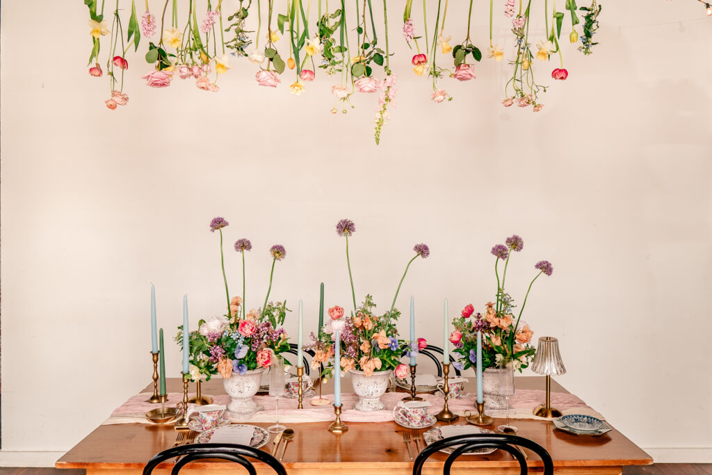 A sweetheart table with playful floral arrangements, blue taper candles, teacups, and flowers hanging from above