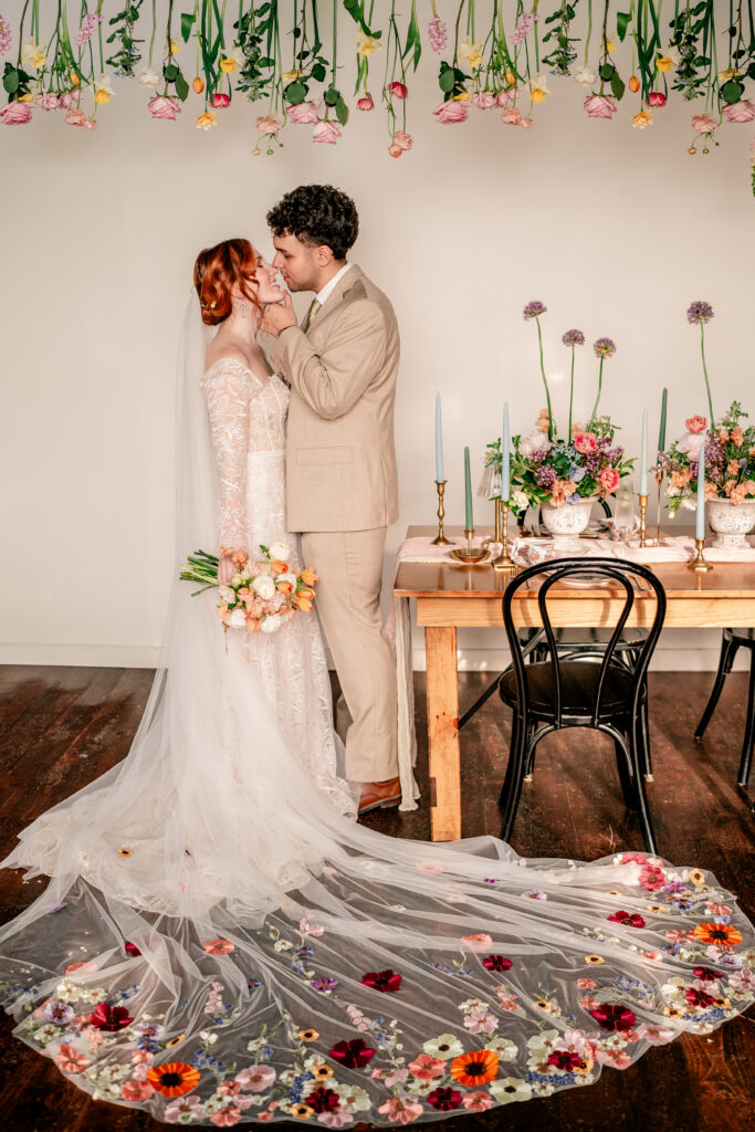 A bride and groom share a kiss beside their sweetheart table, with flowers hanging from the ceiling above