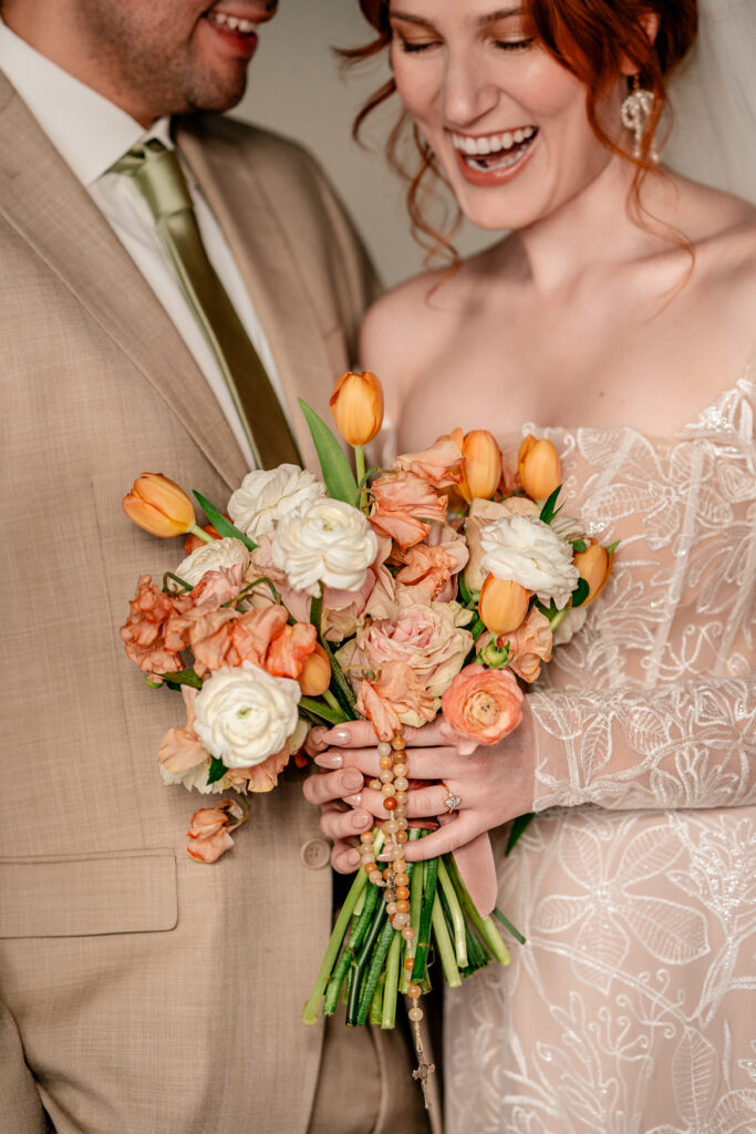 A bride laughing as she holds a bouquet of warm toned flowers and a natural stone rosary for a tea party wedding