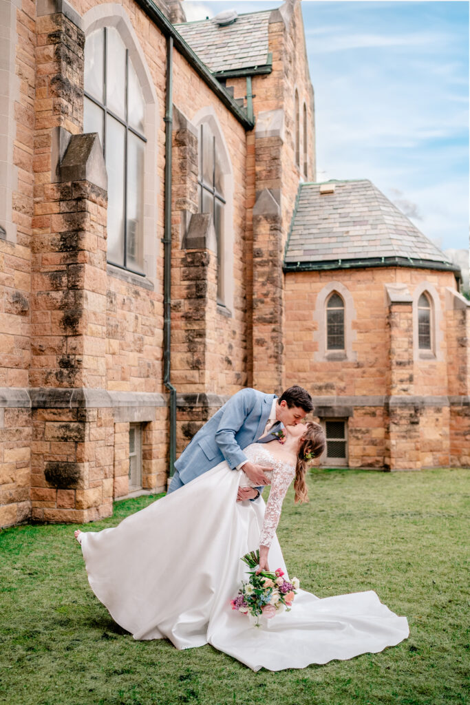 A bride and groom sharing a dip kiss next to their church