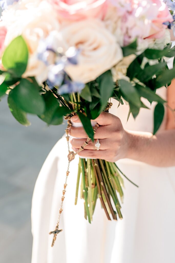 A close up of a bride's wedding ring set on her finger as she holds a pink and blue bouquet of flowers with a rosary hanging from the stems
