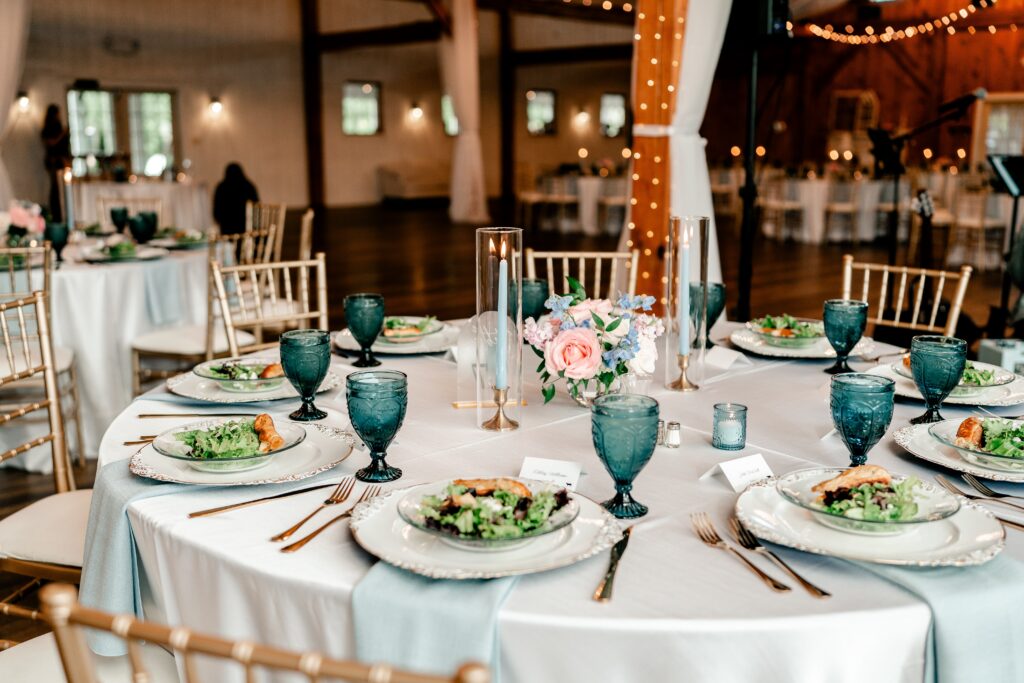 A table set with pink and blue flowers, blue water goblets and napkins, and salads resting on rustic white chargers for a Purcellville wedding photographer